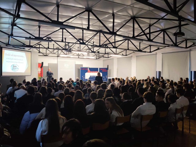 aula szkolna uczniowie siedzą na scenie przemawia do nich mężczyzna kobieta stoi miedzy uczniami koło sceny rzytnik z logo szkoły 