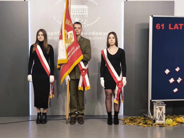 dwie dziewczyny stoją miedzy nimi stoi chłopak ze sztandarem szkolnym za nimi tablica z logo szkoły po prawej stronie granatowa tablica
