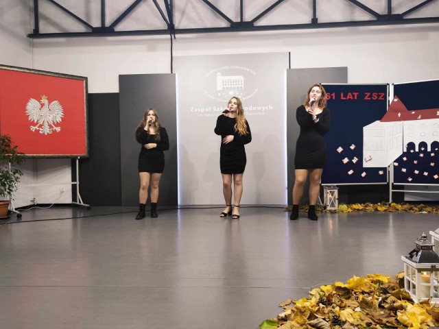 trzy dziewczyny w czarnych sukienkach stoja na scenie i śpiewają po lewej herb Polski w tle szara tablica z logo szkoły