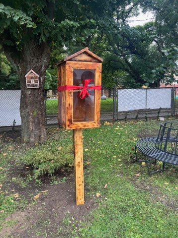 drewniana biblioteczka plenerowa z czerwoną kokardą postawiona na trawniku w tle drzewa